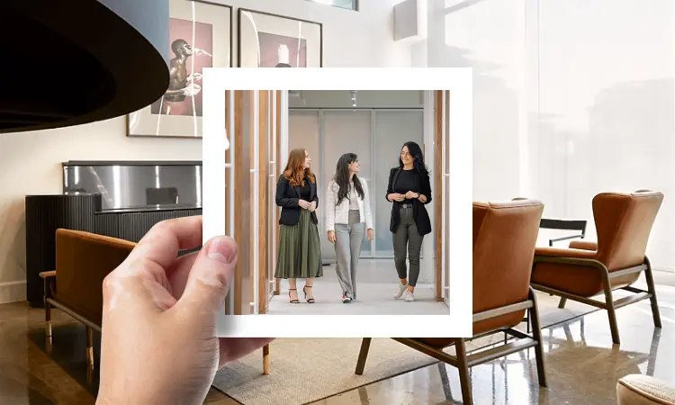 A hand holding up a photo of three colleagues walking through a modern office, framed against the office lounge